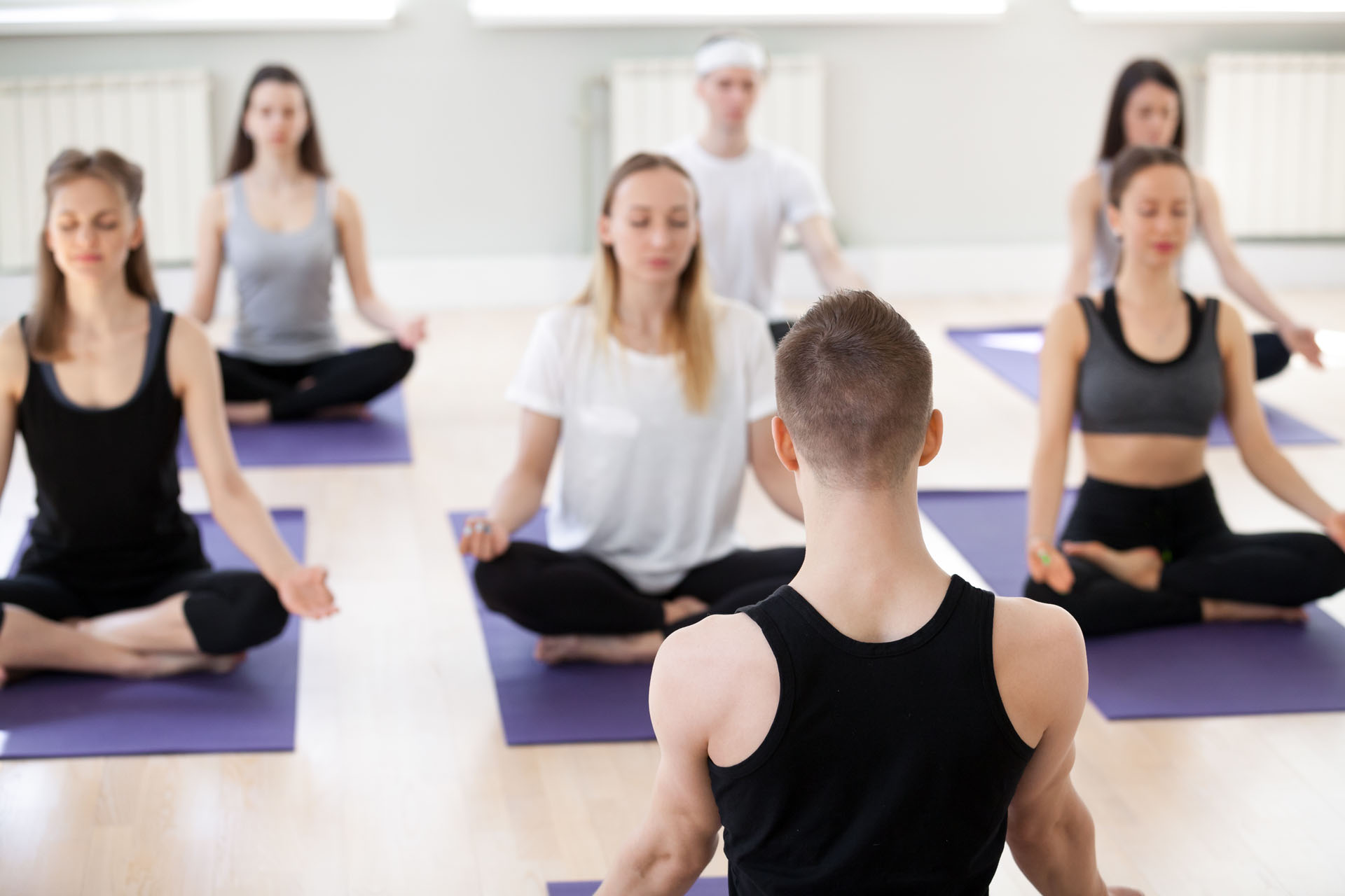 Group of young sporty people practicing yoga lesson with male instructor, doing Easy Seat exercise, Sukhasana pose, working out, indoor session, students training in sport club, teacher rear view
